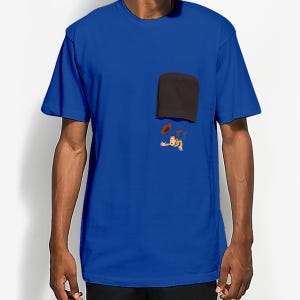 May include: Royal blue t-shirt with a black pocket and a cartoon graphic of a character with a cowboy hat and a red object. The graphic is located below the pocket.