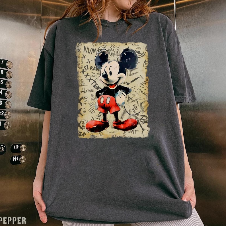 Może przedstawiać: Ciemnoszara koszulka z nadrukiem Mickey Mouse w stylu vintage. Myszka Miki jest przedstawiona w swoim klasycznym stroju z czerwonymi spodenkami i butami, czarną koszulą i białymi rękawiczkami. Tło zawiera tekst w stylu graffiti.