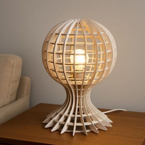 May include: A white, spherical wooden table lamp with a lattice design. The lamp has a bulb inside, and a unique, layered base. The lamp is on a wooden table.