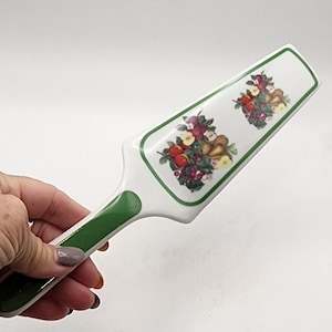 May include: White ceramic cake server with a green handle and trim. The server features two decorative fruit arrangements with red apples, pears, and purple grapes. A classic kitchen utensil for serving cakes and pies.