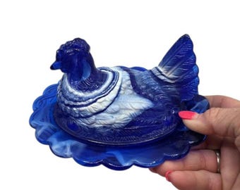 May include: A cobalt blue and white slag glass hen on a nest dish. The piece features a molded chicken figure with detailed feather textures resting on a scalloped oval base. The glass displays a marbled swirl pattern of deep blue and opaque white.