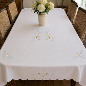 May include: A rectangular white tablecloth featuring a scalloped edge and delicate floral embroidery. The design includes small clusters of pale yellow flowers with light blue stems and leaves positioned at the corners and along the sides.