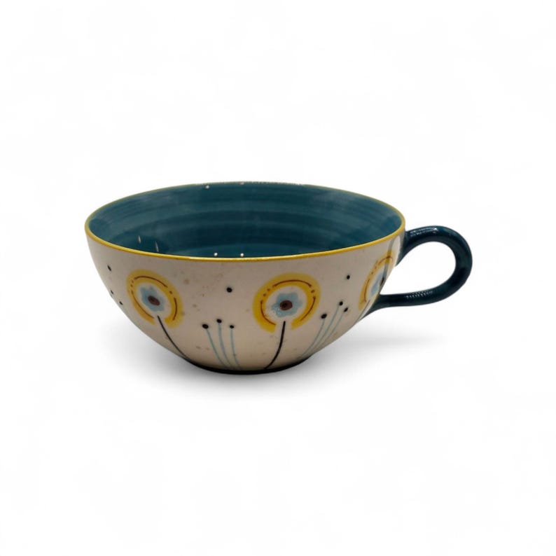 May include: A ceramic teacup with a wide, rounded bowl and a dark blue handle. The interior is dark blue, and the exterior is off-white with yellow and blue floral designs. A thin yellow line accents the rim.