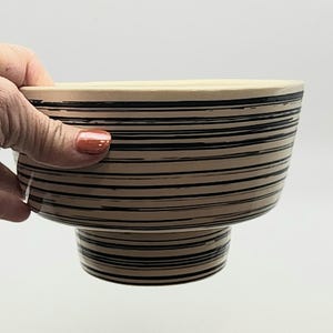 May include: A beige and black striped ceramic bowl with a pedestal base. The bowl has a wide, open top and is held by a hand. The black stripes are horizontal and evenly spaced, creating a textured effect. The bowl's interior is a lighter beige.