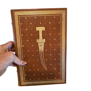 May include: A rectangular brown leather-bound book featuring a gold-stamped dagger illustration centered on the front cover. The cover is decorated with a repeating pattern of small gold crosses and a double-line gold border frame.