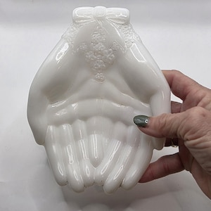 May include: A white, hand-shaped decorative dish. The dish features a detailed design of two hands cupped together, with a bow and floral accents at the top. The dish is made of a glossy, opaque material.