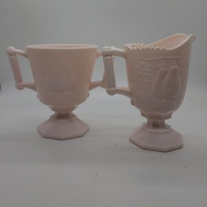 K&ouml;nnte beinhalten: Ein Paar hellrosa Glasservierschalen. Eine ist eine Zuckerdose mit Griffen und Deckel. Die andere ist ein Sahnek&auml;nnchen mit einem Birnenmuster. Beide haben einen Sockel und sind wahrscheinlich Vintage.