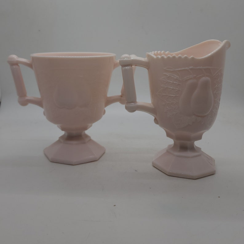K&ouml;nnte beinhalten: Ein Paar hellrosa Glasservierschalen. Eine ist eine Zuckerdose mit Griffen und Deckel. Die andere ist ein Sahnek&auml;nnchen mit einem Birnenmuster. Beide haben einen Sockel und sind wahrscheinlich Vintage.