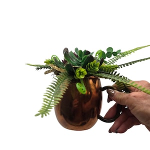 May include: A metallic copper-colored mug filled with an arrangement of artificial greenery, including various shades of green succulent plants, fern fronds, and small sprigs of eucalyptus leaves, creating a dense botanical display.