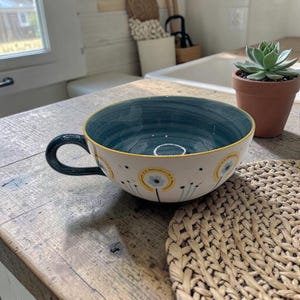 May include: A large ceramic bowl with a dark teal interior and a white exterior decorated with yellow and blue floral designs. The bowl has a dark teal handle and a yellow rim. The bowl is on a wooden surface.