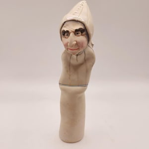 May include: A ceramic figurine of a character with a white hooded hat and a long, curved nose. The figure has a pale complexion with painted facial features, including dark eyebrows and red lips. The body is cylindrical with a blue and white band.