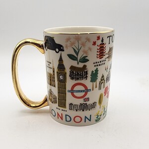 May include: A cream-colored ceramic mug with a gold handle and rim. The mug is decorated with illustrations of London landmarks and Japanese imagery. The word "LONDON" is printed in blue at the bottom of the mug.