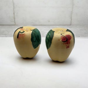 May include: A pair of ceramic salt and pepper shakers shaped like apples. The shakers are cream-colored with green leaves and the letters "P" and "S" painted on them. The apple-shaped shakers have small holes on top for dispensing salt and pepper.