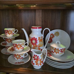 May include: A white ceramic tea set featuring a vibrant orange, red, and yellow floral pattern. The set includes a tall teapot with a red lid, a creamer, a lidded sugar bowl, four teacups with matching saucers, and several stacked plates.