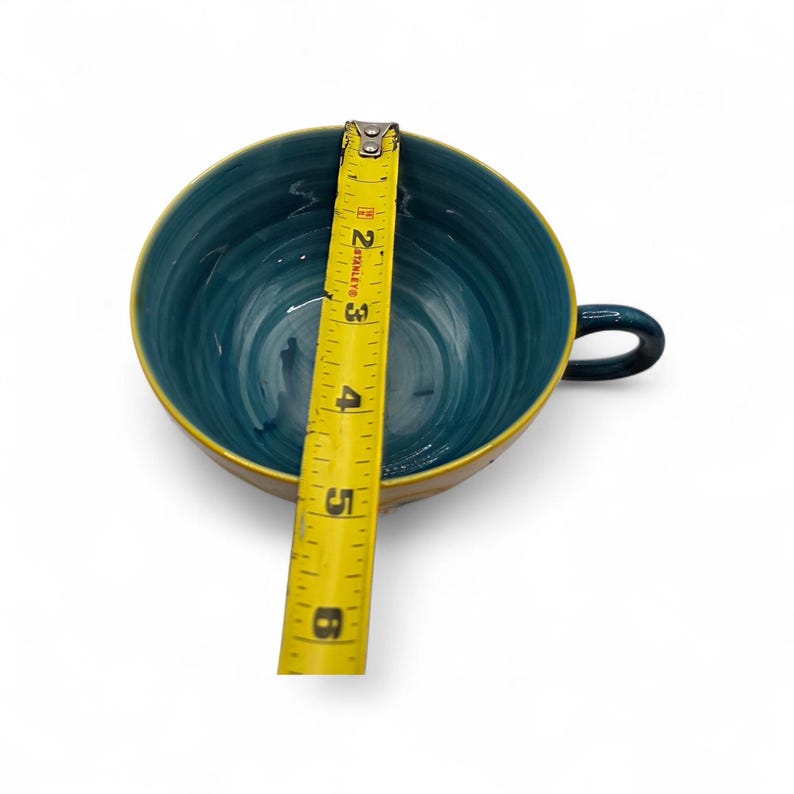 May include: A teal and yellow ceramic teacup with a handle, measuring approximately 5 inches in diameter. A yellow measuring tape is placed inside the cup, showing the diameter. The cup has a glossy finish and a simple design.