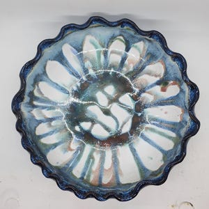 May include: A handmade ceramic bowl with a unique, scalloped edge. The bowl features a striking design with shades of blue, white, and brown, creating a floral pattern. The rim is a darker blue, adding contrast to the overall design.