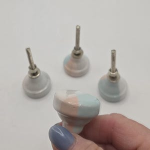 May include: Four ceramic cabinet knobs with a pastel color scheme. The knobs have a rounded shape with a metal screw. The colors include light blue, pink, and white. The knobs are for home decor.