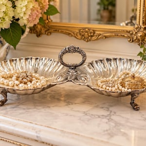 May include: A silver-toned metal double-bowl serving dish with a central ornate handle and scalloped edges. The dish features four small decorative feet and contains strands of white pearl necklaces and small gold-toned trinket boxes.