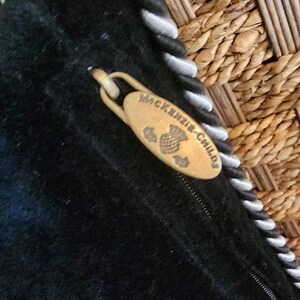 Op de afbeelding: Close-up van een zwart stoffen item met een goudkleurige ritstrekker. De ritstrekker is ovaal van vorm en heeft de woorden "MacKenzie-Childs" en een distelontwerp gegraveerd. De rand van het item heeft een wit en zwart touwachtig randje.