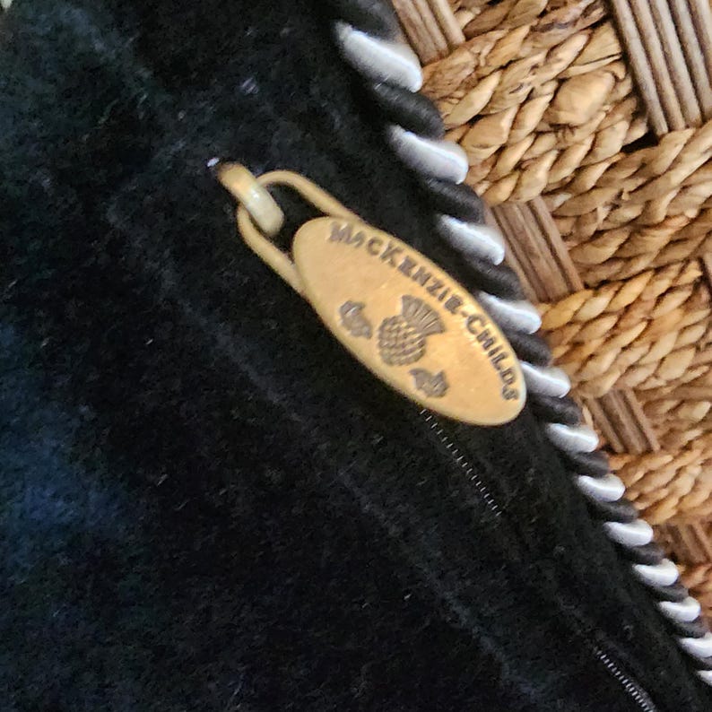 Op de afbeelding: Close-up van een zwart stoffen item met een goudkleurige ritstrekker. De ritstrekker is ovaal van vorm en heeft de woorden "MacKenzie-Childs" en een distelontwerp gegraveerd. De rand van het item heeft een wit en zwart touwachtig randje.