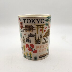 May include: A white ceramic travel mug with a gold rim, featuring colorful illustrations of landmarks and the city names "TOKYO" and "MEXICO CITY". The design includes pagodas, temples, and other architectural elements.