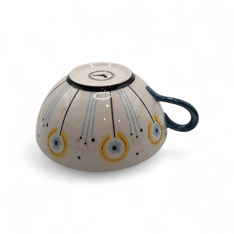 May include: A ceramic teacup with a cream-colored exterior, adorned with blue and yellow floral patterns and black accents. The handle is a dark teal color. The cup is approximately 4 inches in diameter and 3 inches tall.