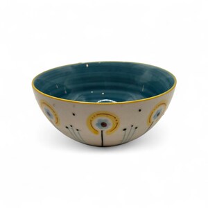 May include: A ceramic bowl with a cream-colored exterior and a teal interior. The bowl is decorated with yellow and blue floral designs. The rim is outlined in yellow. The bowl is likely used for serving food or as a decorative piece.