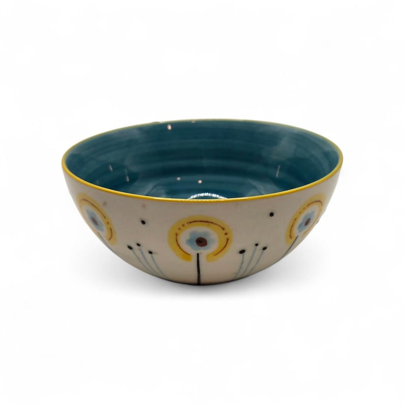 May include: A ceramic bowl with a cream-colored exterior and a teal interior. The bowl is decorated with yellow and blue floral designs. The rim is outlined in yellow. The bowl is likely used for serving food or as a decorative piece.