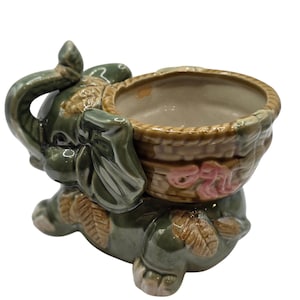 May include: A glossy ceramic elephant figurine in a dark olive green glaze, featuring a brown woven-style basket on its back. The basket has a light pink decorative accent on the side, and the elephant has brown leaf-shaped details on its legs.