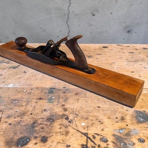 May include: A vintage wooden hand plane with a dark wooden handle and a metal blade. The plane has a rectangular wooden base and a curved handle. The metal blade is visible at the top of the plane. The tool is designed for woodworking.