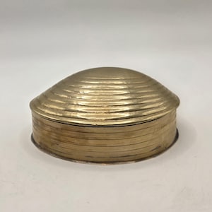 May include: A round brass box with a domed lid featuring concentric circular ridges. The base of the container also displays horizontal linear detailing around its circumference, creating a consistent metallic texture across the entire object.