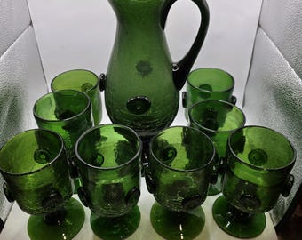 May include: A green glass pitcher with a black handle and a set of matching green glasses. The pitcher and glasses have a textured surface and a dark green color. The set includes a pitcher and eight glasses.