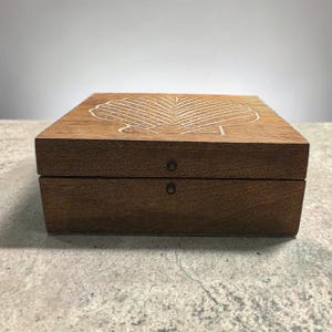 May include: A square, wooden box with a hinged lid. The box is a warm brown color and features a carved seashell design on the top. Two small, round metal hinges secure the lid. The box is sitting on a gray surface.