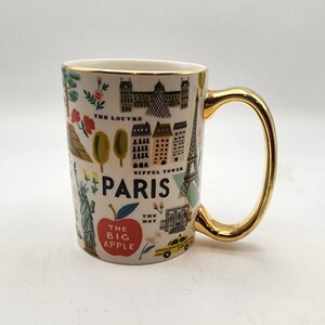 May include: White ceramic mug with a gold handle and rim, featuring illustrations of Parisian landmarks like the Eiffel Tower and the Louvre. The mug also includes the text "PARIS" and "The Big Apple."