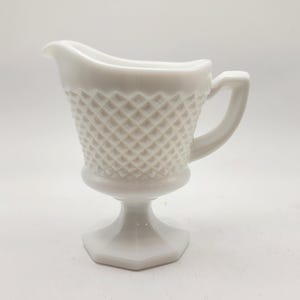 May include: A white, vintage creamer with a diamond pattern. The creamer has a handle and a spout for easy pouring. It sits on a pedestal base with a hexagonal shape. The creamer is made of a solid, opaque material.