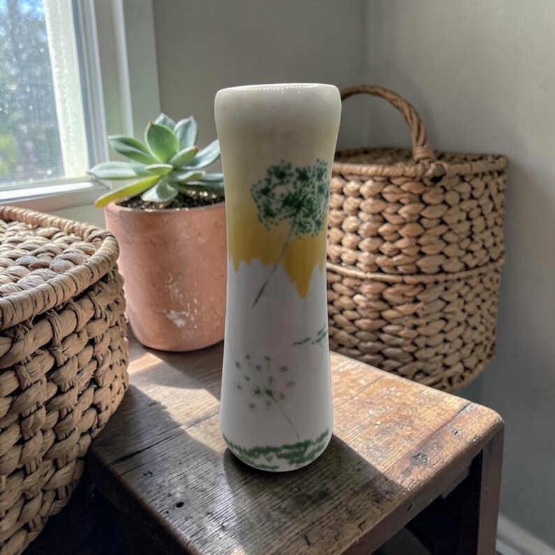 May include: A white ceramic vase with a yellow and green dandelion design. The vase is cylindrical with a slightly flared top. The dandelion design is painted in green and yellow. The vase is on a wooden surface.