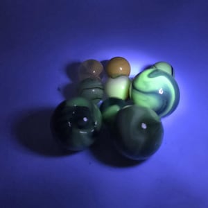 May include: A collection of glass marbles in various colors, including green, yellow, and brown. The marbles have a swirled or speckled design. The image is taken against a blue background.