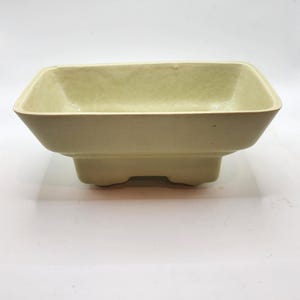 May include: A rectangular, light green ceramic planter with a slightly textured surface. The planter has a raised base with three small feet. The interior is a lighter shade of green. The planter is designed for small plants or succulents.