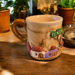 May include: Hand-painted ceramic mug with a brown handle and a village scene. The mug features a tan base color with a village scene in shades of red, purple, green, and white. The mug is sitting on a wooden surface.