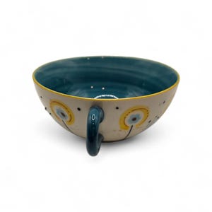 May include: A ceramic teacup with a teal interior and a cream-colored exterior. The cup features a yellow rim and a teal handle. The exterior is decorated with yellow and blue floral designs and black dots.