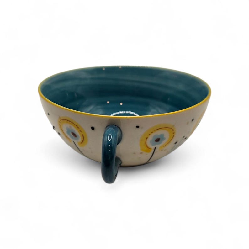 May include: A ceramic teacup with a teal interior and a cream-colored exterior. The cup features a yellow rim and a teal handle. The exterior is decorated with yellow and blue floral designs and black dots.