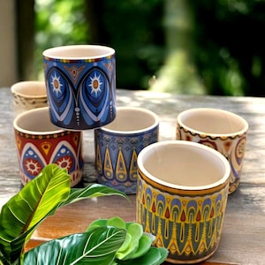 May include: Six ceramic mugs with vibrant, patterned designs. The mugs display patterns in blue, red, yellow, and beige. They are arranged on a wooden surface, with green foliage in the foreground. Each mug has a unique design.