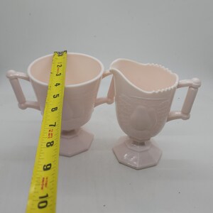 K&ouml;nnte beinhalten: Ein hellrosa Zuckerdosen- und Sahne-Set. Die Zuckerdose ist etwa 10 cm hoch und 10 cm breit. Der Sahneaufgie&szlig;er hat ein Birnenmuster und ist etwa 10 cm hoch. Beide Teile haben Griffe und stehen auf einem sechseckigen Sockel.