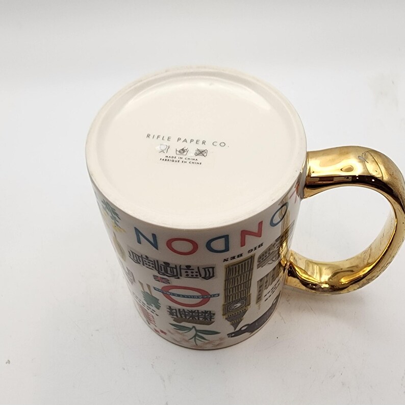 May include: A ceramic mug with a gold-colored handle and a cream-colored body. The mug is decorated with illustrations of London landmarks and the word "LONDON" in large letters. The bottom of the mug has the text "RIFLE PAPER CO."