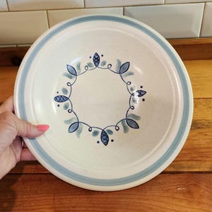 May include: Cream-colored ceramic plate with a blue floral design. The plate has a light blue border and a central pattern of blue leaves and swirls. The plate is on a wooden surface.