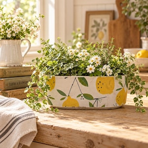 May include: An oval ceramic planter with a white base featuring a hand-painted pattern of yellow lemons, green leaves, and black stems. The planter is filled with a variety of small green plants and white daisy-like flowers, creating a lush display.