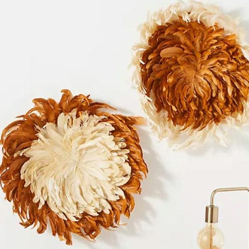 May include: Two decorative wall hangings made of feathers. The circular pieces feature a mix of brown and cream-colored feathers, creating a layered, textured effect. The wall hangings are mounted on a white wall, adding a touch of bohemian style.