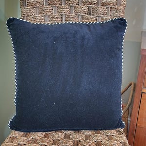 Op de afbeelding: Een vierkant, marineblauw kussen met een wit en marineblauw koord. Het kussen is gemaakt van een zacht, fluwelig materiaal en staat op een gevlochten stoel.