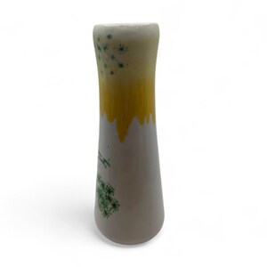 May include: A slender, off-white ceramic vase with a yellow band that drips down the sides. Green floral designs are painted on the lower portion of the vase. The vase has a slightly flared top and a smooth, glossy finish.