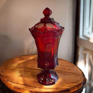 May include: A ruby red glass jar with a lid, standing on a wooden surface. The jar has a pedestal base and a decorative lid with circular accents. The glass has a faceted design, adding to its vintage appeal. The jar is likely used for storage or display.